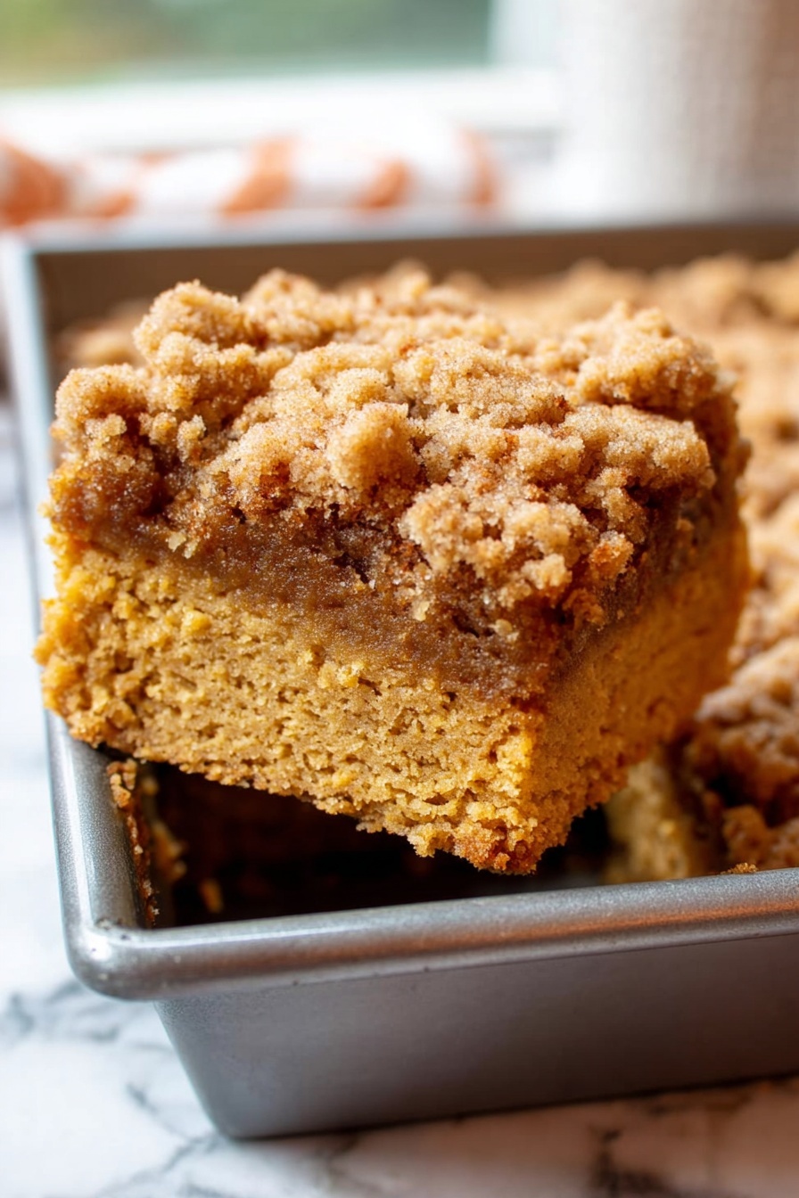 Two thick square slices of crumb cake are stacked on a white plate, placed on a white marbled surface. Each slice shows two layers: a dense, moist bottom layer with a rich orange-brown color and a lighter, textured crumbly top layer that is slightly uneven and golden brown. Crumbs are scattered around the plate, adding to the rustic look. In the background, there is a blurred glimpse of a baking pan and a leafy green view outside a window. The photo taken with an iphone --ar 2:3 --v 7