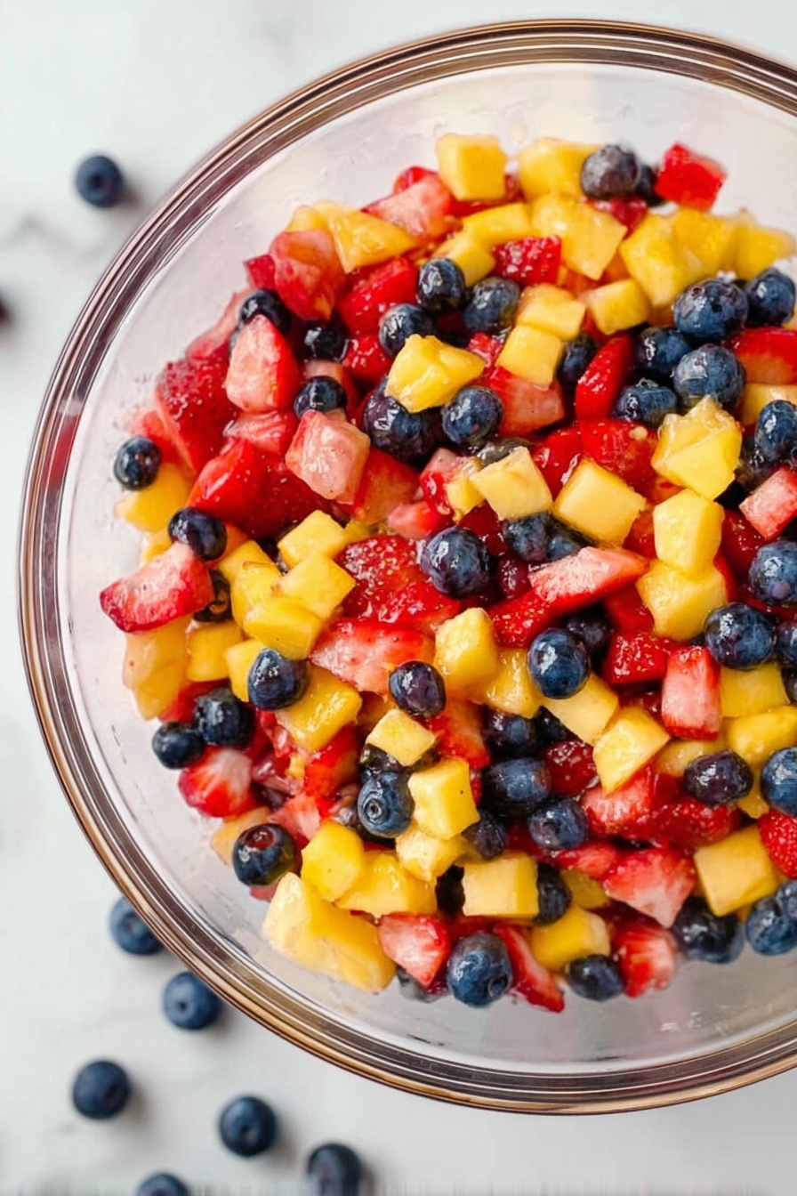 A clear glass bowl filled with a colorful fruit salad, showing three main layers of small, chopped fruits: bright yellow mango pieces, red strawberries, and dark blue blueberries scattered throughout. The textures vary with smooth blueberries rolling around, soft cubed strawberries, and juicy mango bits, all mixed together. The bowl is set on a white marbled surface with a few blueberries resting next to it. The bright colors of the fruits contrast well with the clear bowl and the pale background. Photo taken with an iphone --ar 2:3 --v 7