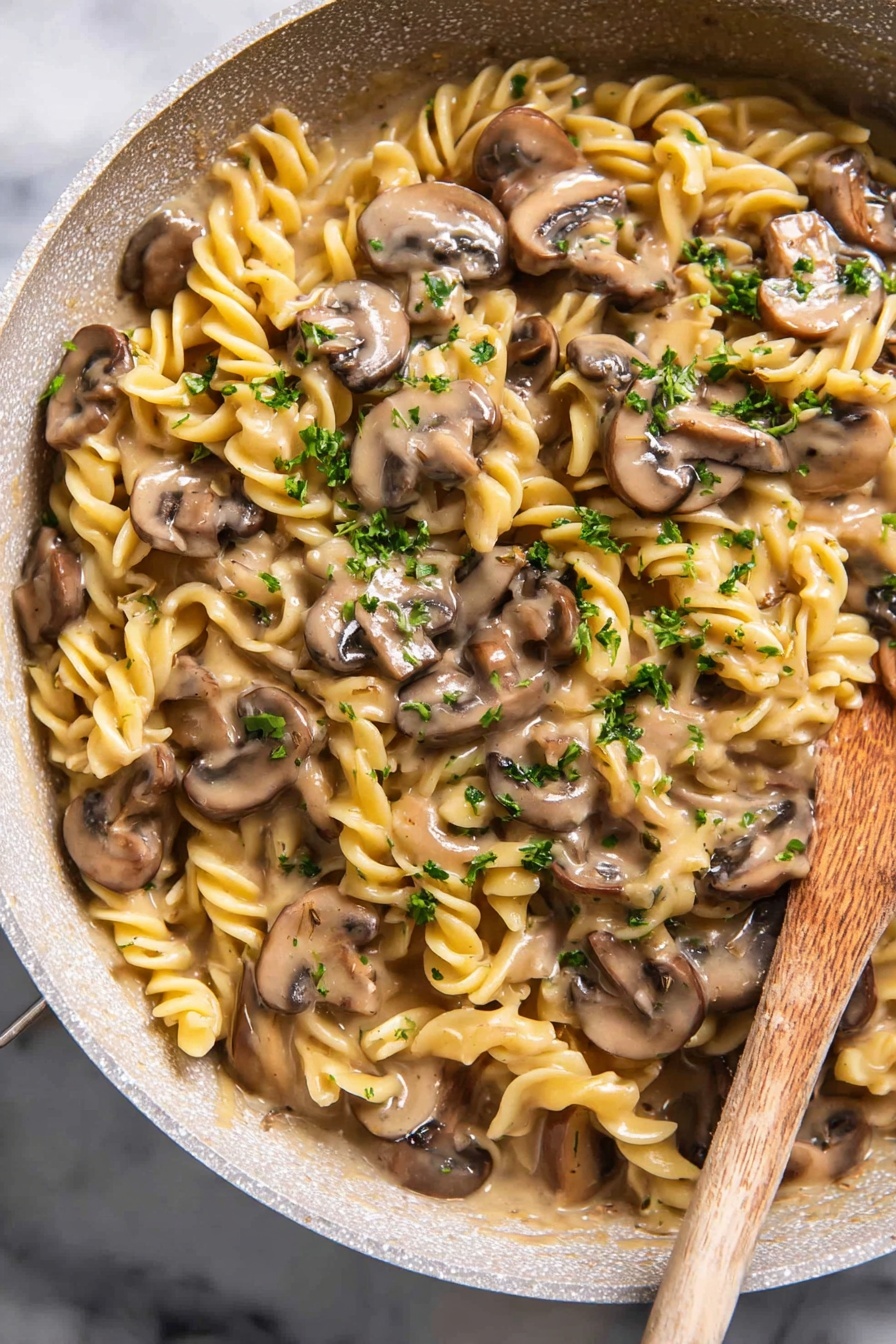 Vegan Mushroom Stroganoff - The image shows a round light beige pot filled with creamy rotini pasta and sliced mushrooms mixed together. The twisted pasta is pale yellow while the mushrooms are medium brown with lighter edges. Small pieces of green parsley are sprinkled over the pasta. A wooden spoon is inside the pot, partially covered with the pasta dish. The pot is placed on a white marbled surface with two whole mushrooms seen in the top left corner. Photo taken with an iphone --ar 2:3 --v 7