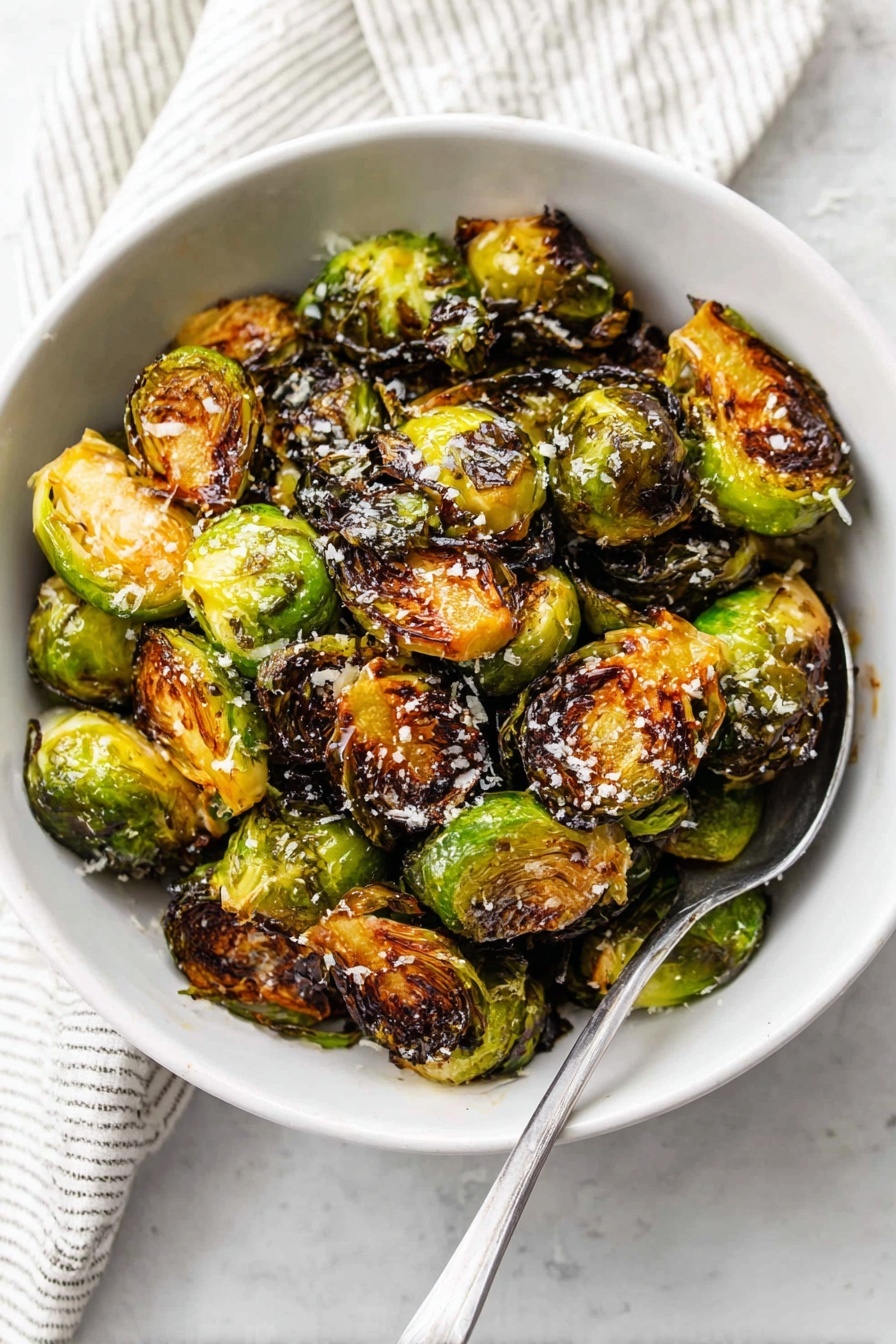 A white bowl filled with one layer of roasted Brussels sprouts, each halved, showing a mix of green, golden brown, and charred dark brown colors with a slightly crispy texture. Some of the Brussels sprouts have a light sprinkle of white grated cheese on top. A silver spoon rests on the right side inside the bowl. The bowl sits on a white marbled surface with part of a striped cloth visible at the top. Photo taken with an iphone --ar 2:3 --v 7