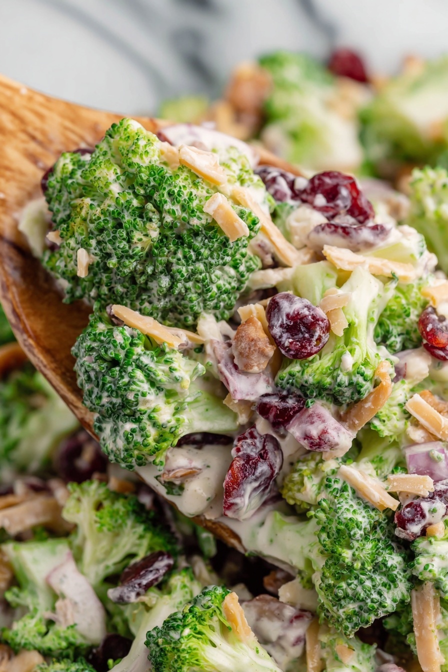 A clear glass bowl filled with a broccoli salad made up of many green broccoli florets with a creamy white dressing mixed over them. Small pieces of light brown bacon, thin orange shredded cheese, dark red dried cranberries, and tiny beige sunflower seeds are scattered throughout the salad, adding texture and color. The bowl is placed on a white marbled surface, with a small white bowl of dried cranberries and a silver pepper grinder in the background. Photo taken with an iphone --ar 2:3 --v 7