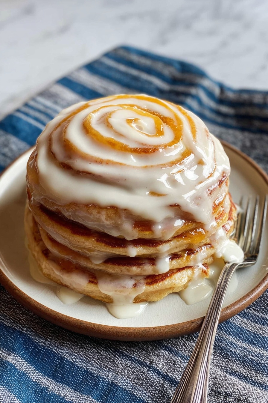 Two thick, fluffy pancakes stacked on a round white plate, with a swirl of amber syrup on top, glistening and slightly dripping down the sides. A silver fork rests beside the stack. In the background, slightly blurred, is a small white bowl filled with a creamy, pale spread. The scene is set on a white marbled surface with a blue cloth napkin partially visible. photo taken with an iphone --ar 2:3 --v 7