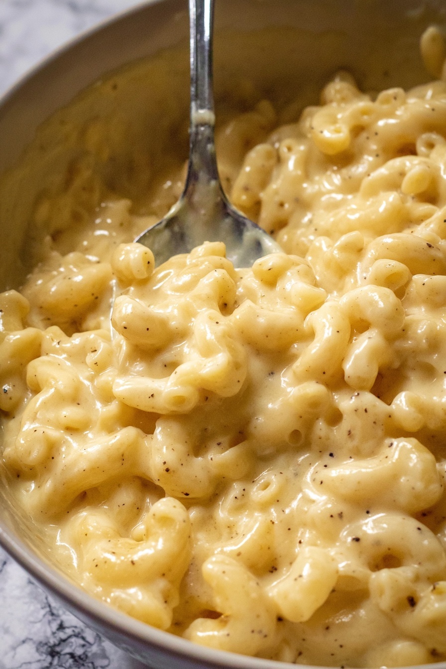 A close-up view of creamy macaroni and cheese being lifted by a metal spoon from a pot. The dish has a thick, smooth yellow cheese sauce that fully covers the elbow-shaped pasta. The cheese sauce has a rich, glossy texture and small specks of seasoning can be seen throughout. The background shows more macaroni and cheese in the pot with the same creamy and uniform texture. photo taken with an iphone --ar 2:3 --v 7