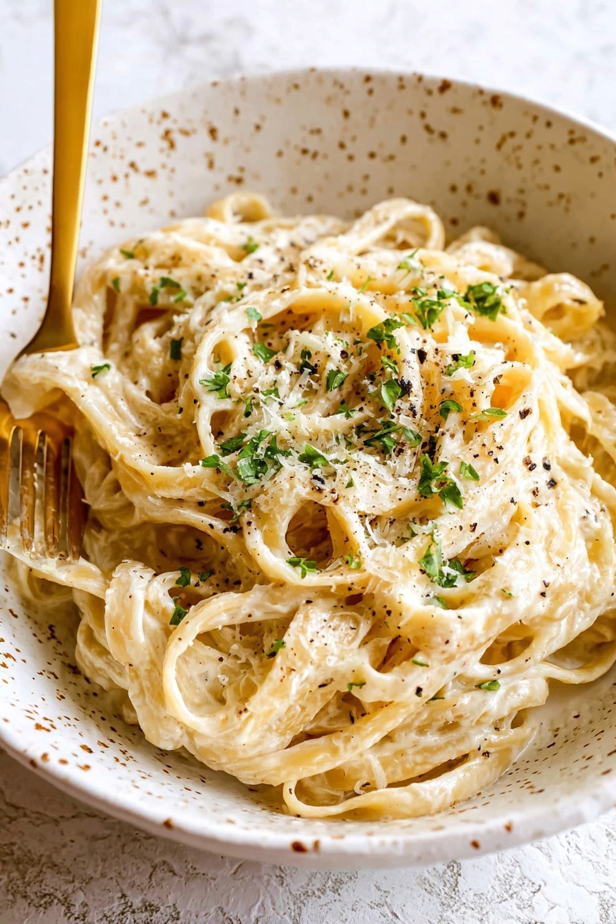 A close-up view of creamy fettuccine pasta piled high on a white plate, with the noodles covered in a smooth, pale yellow sauce. The noodle strands have a soft, slightly twisted texture and are sprinkled evenly with small bits of green parsley and black pepper. A gold spoon with a dark handle is placed near the edge of the plate, partially buried in the pasta. The background is a white marbled surface. photo taken with an iphone --ar 2:3 --v 7