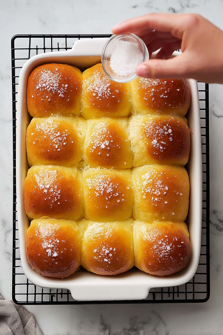 A shiny golden brown bread roll with a soft, fluffy inside sits on a simple white plate in the front. The top of the roll is sprinkled with coarse salt crystals that catch the light. Behind it is a white baking dish full of similarly golden rolls, close together with a slightly glossy finish. To the right, on the white marbled surface, is a small white bowl holding two pale yellow cubes of butter. The whole scene is bright with soft light, showing the warm colors and textures clearly. photo taken with an iphone --ar 2:3 --v 7