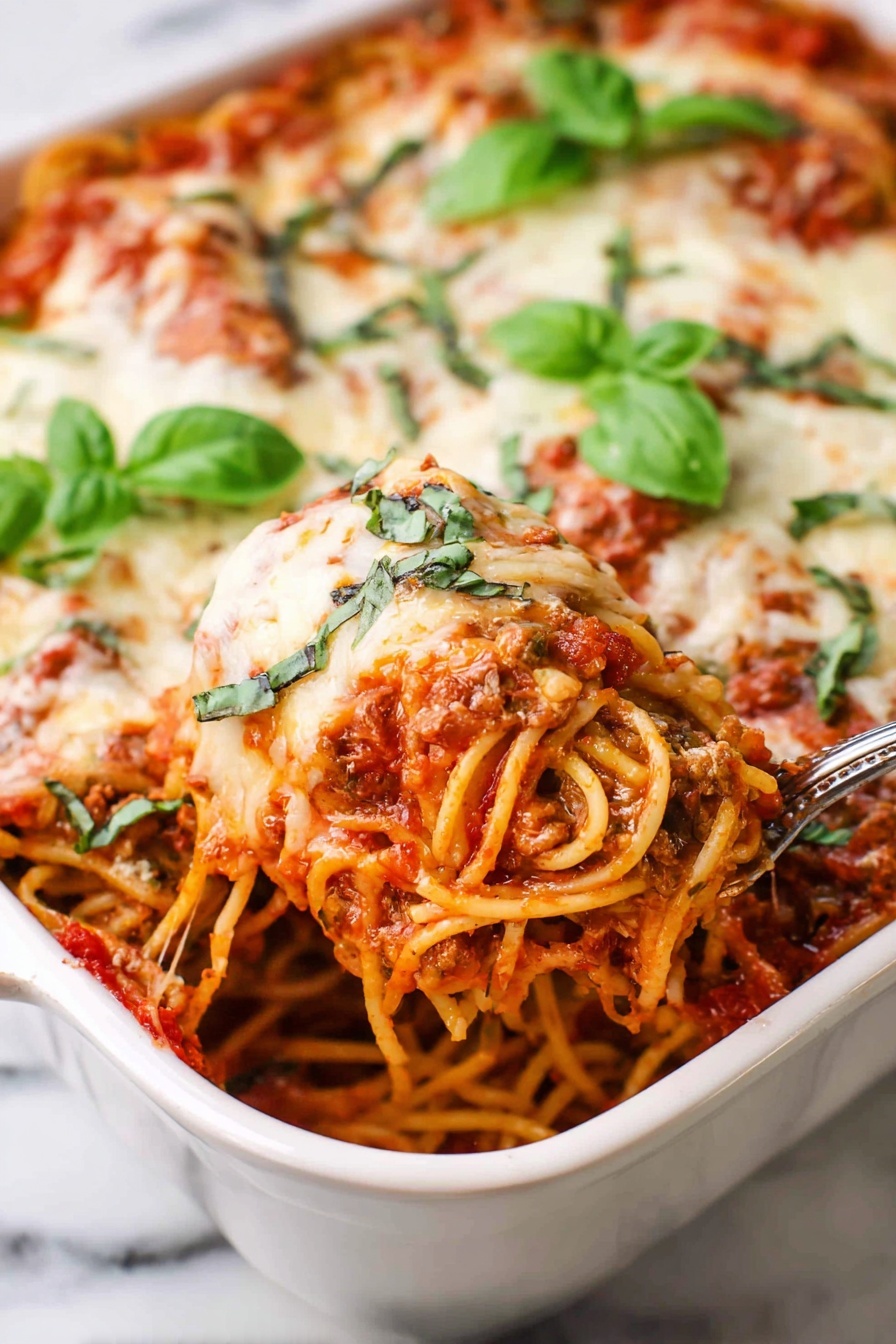 The dish shows a close-up of baked spaghetti layered in a white ceramic baking dish, with a base layer of red tomato sauce mixed with bits of meat and herbs, followed by a thick layer of spaghetti noodles coated in the sauce. On top, there is a golden melted layer of cheese with green chopped basil scattered all over, and a few whole fresh basil leaves placed on top as garnish. A spoon is scooping out a portion of the spaghetti, revealing the rich, saucy texture and strands of noodles beneath the cheese. The whole dish sits on a surface with a white marbled texture. photo taken with an iphone --ar 2:3 --v 7
