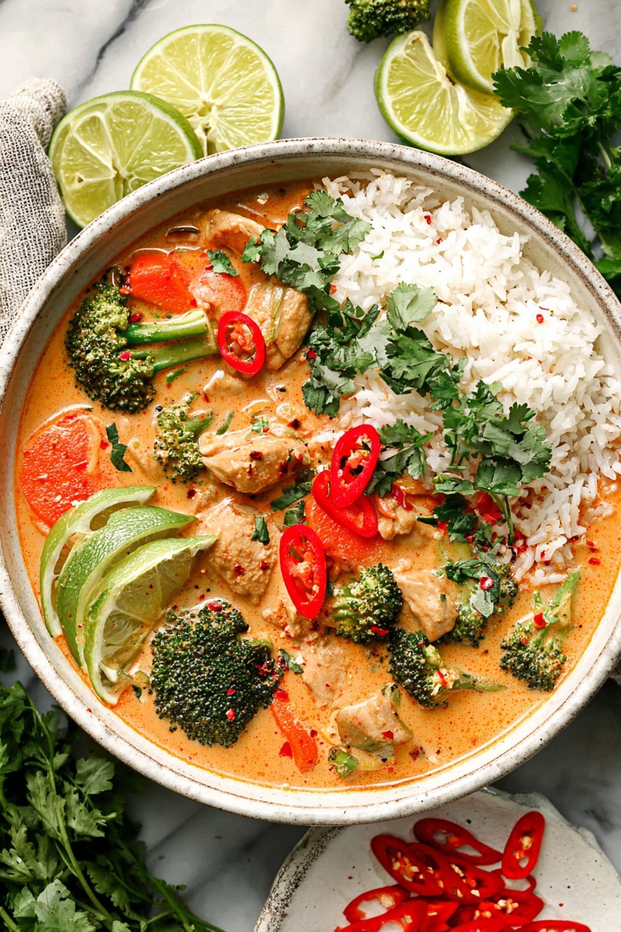 A bowl filled with a creamy orange curry soup that contains pieces of chicken, broccoli florets, and sliced carrots, all mixed together. On one side of the bowl, there is a layer of white rice topped with green cilantro leaves. There are thin red chili slices scattered on the soup, adding bright red spots. Inside the bowl, there are three lime wedges sitting on top of the rice on the left side. A vintage gray spoon is placed on the right side of the bowl. The bowl is white and sits on a white marbled surface, surrounded by halved limes and a small white plate with sliced red chili peppers. The dish looks fresh with bright colors of green, orange, red, and white. Photo taken with an iphone --ar 2:3 --v 7