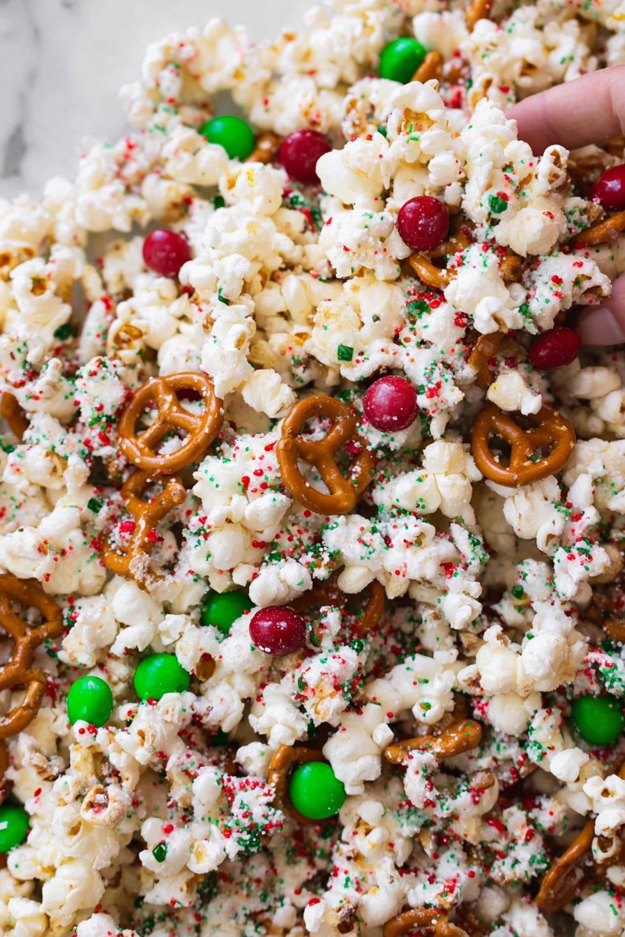 The image shows a close-up view of a festive popcorn mix. The base layer is white popcorn with a light, fluffy texture. Scattered throughout are small, round pretzels with a glossy, brown surface. Mixed in are red and green candy-coated chocolates, adding bright pops of color. The entire mix is sprinkled with tiny red, green, and white sugar crystals, giving a textured and colorful look. The background is a white marbled surface. photo taken with an iphone --ar 2:3 --v 7