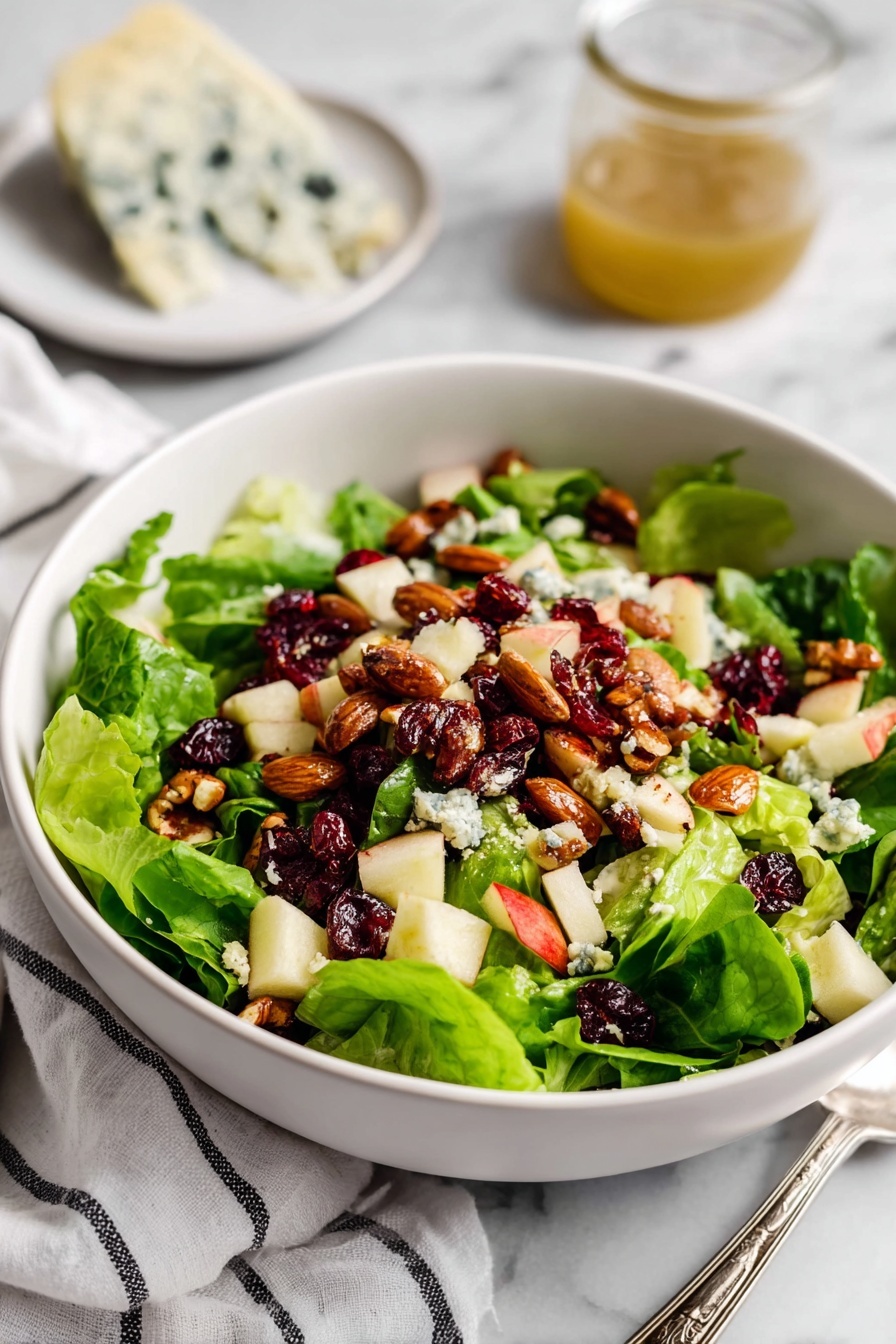 A white bowl filled with a fresh salad showing three main layers: the base layer is bright green leafy lettuce, the middle layer has light cream and red small chopped apple pieces, and the top layer is sprinkled with roasted brown almonds, dark red dried cranberries, and small chunks of pale blue cheese with dark veins. In the background, there is a small white plate with a piece of blue cheese, a silver spoon, and a glass jar with golden dressing on a white marbled surface. A white cloth with thin black stripes is placed near the bowl. Photo taken with an iphone --ar 2:3 --v 7