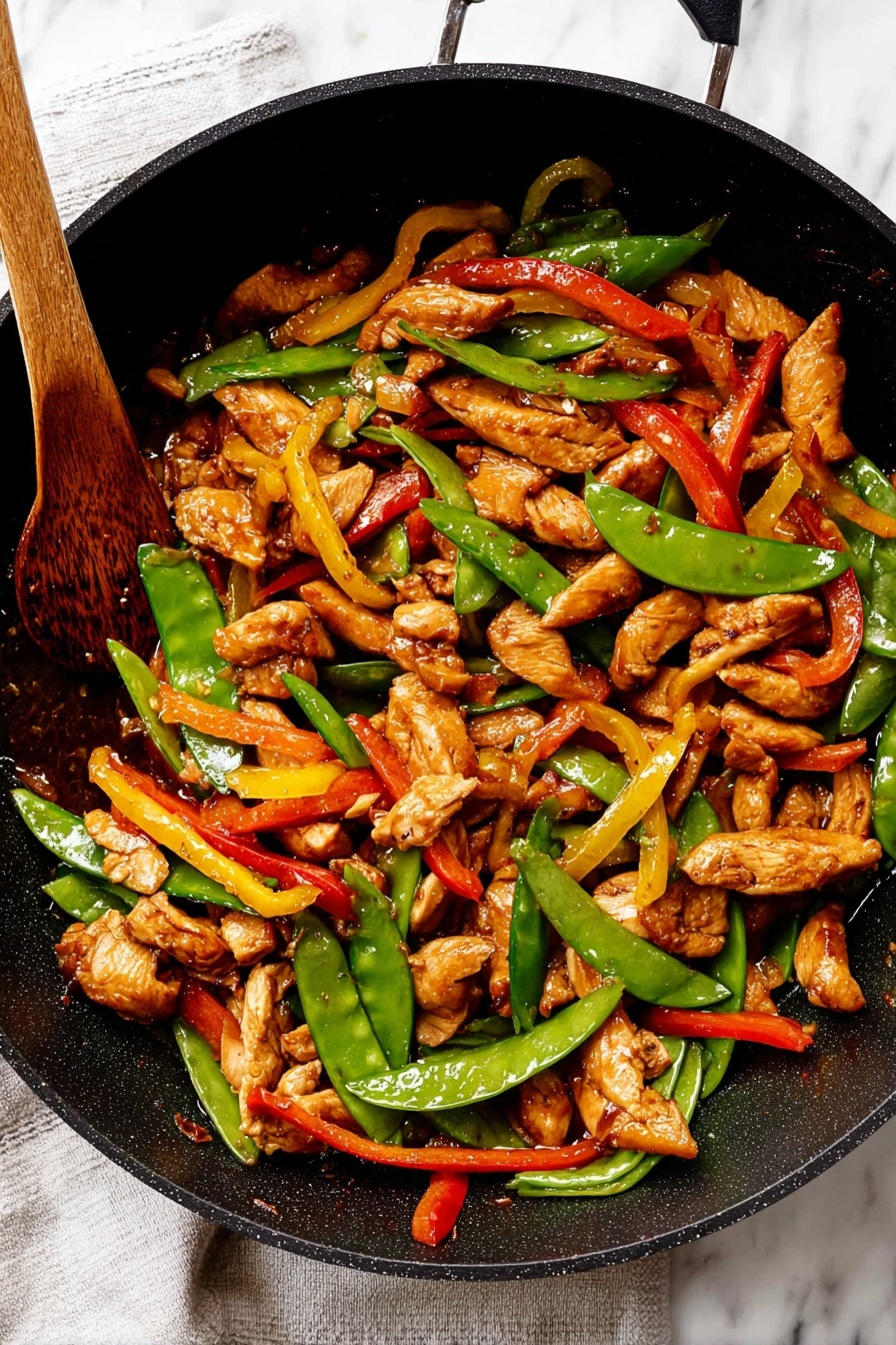 The image shows a black pan filled with stir-fried slices of cooked light brown chicken mix, bright green sugar snap peas, and thin strips of red and yellow bell peppers. The chicken pieces are glossy and scattered evenly throughout the pan, mixed with crisp vegetables that add color contrast. A wooden spoon with some sauce on it is resting inside the pan on the left side. The pan sits on a cloth over a white marbled surface. photo taken with an iphone --ar 2:3 --v 7