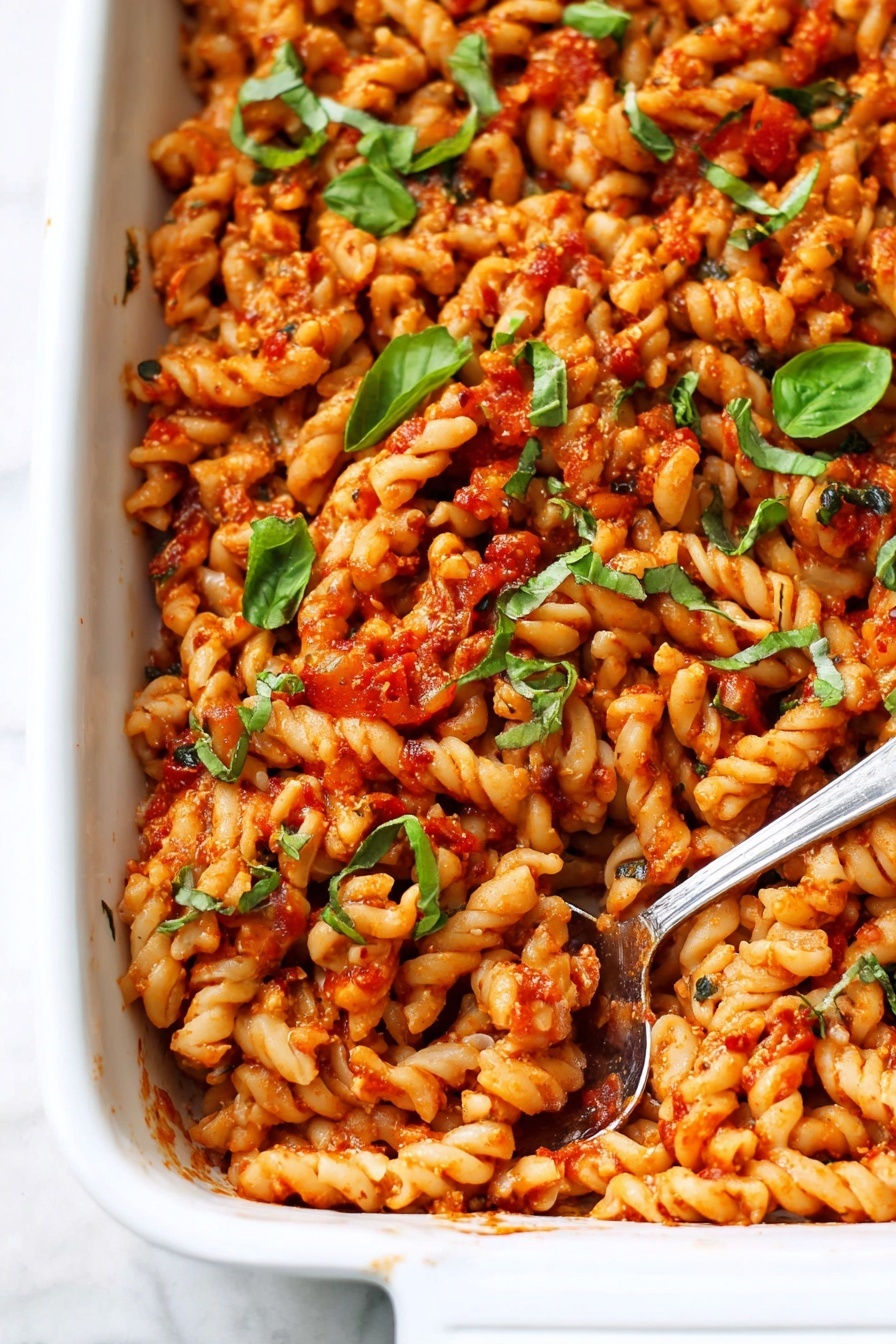 The image shows a white rectangular baking dish filled with a creamy pasta bake. The pasta is twisted, lightly golden in color, and mixed with pieces of sun-dried tomatoes and green herbs, mainly fresh basil leaves scattered on top. The texture looks soft and creamy with small bits of herbs and tomato adding color contrast. A silver serving spoon is slightly dipped into the dish from the right side. The dish sits on a white marbled surface with a few fresh basil leaves scattered around. photo taken with an iphone --ar 2:3 --v 7