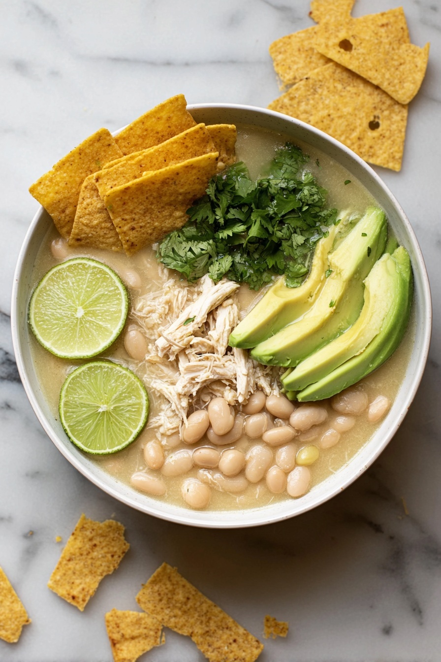 A white bowl filled with a creamy white soup containing shredded white chicken pieces and white beans, topped with a small pile of crispy golden tortilla strips, bright green avocado slices arranged on one side, and small green cilantro leaves scattered on top, all sitting on a white plate that holds three lime wedges and a silver spoon. The background is a white marbled texture. Photo taken with an iphone --ar 2:3 --v 7