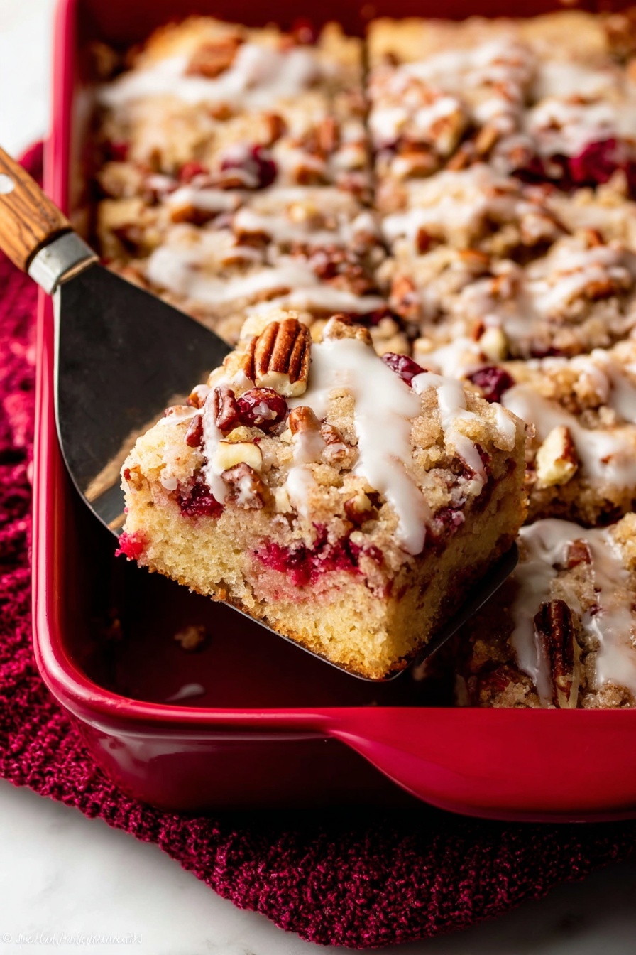 A close-up view of a red baking dish filled with a square crumb cake. The cake has three visible layers: the bottom layer is light yellow sponge with scattered bright red berries, the middle layer has a mix of crunchy light brown crumb topping and pecans, and the top layer shows a light white glaze drizzled over the nuts and crumbs. A metal spatula with a wooden handle lifts one square piece, revealing soft crumb texture and berry bits inside. The dish sits on a white marbled surface with a textured deep red cloth nearby. photo taken with an iphone --ar 2:3 --v 7