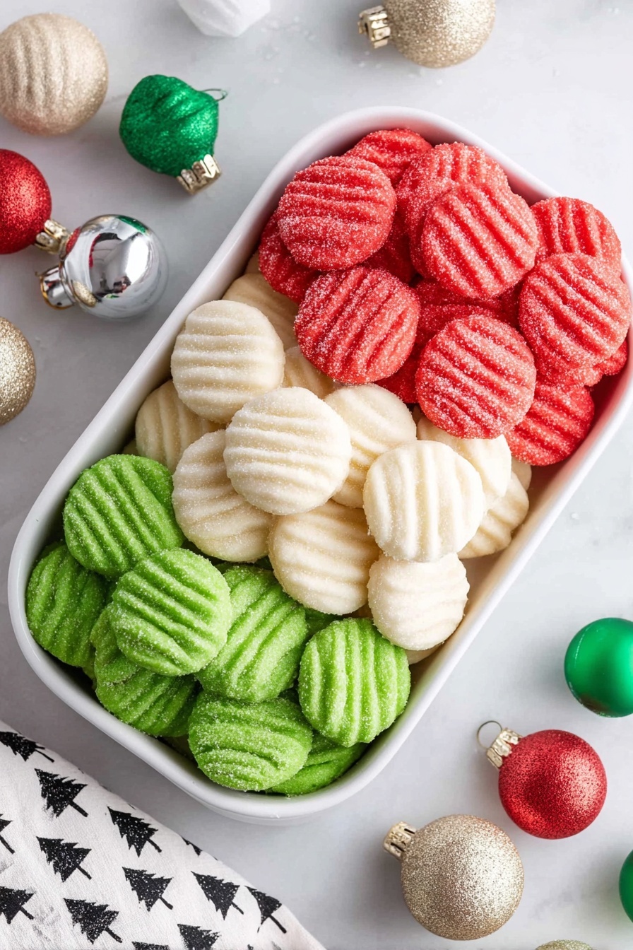 A white divided dish with three sections stacked with small round cookies, each having ridged patterns on top and a light dusting of sugar. The top section holds bright red cookies, the middle section has white cookies, and the bottom section contains vibrant green cookies. Around the dish, there is a white marbled surface decorated with small festive bells in red, green, and cream colors, and glittery holiday ornaments in gold, silver, white, and green. A white cloth with black tree patterns is partially visible in the corner. photo taken with an iphone --ar 2:3 --v 7