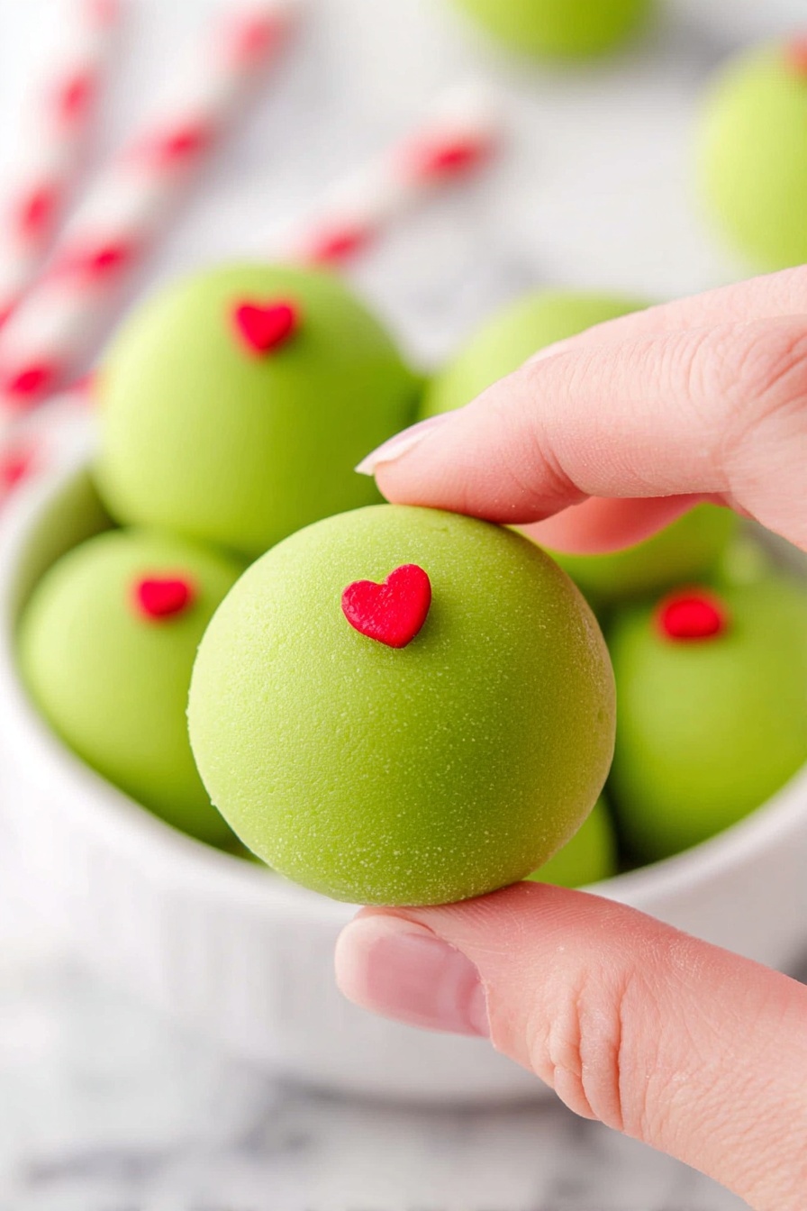 A close-up image of a light green round dessert with a matte smooth texture, topped with a small red heart shaped decoration in the center. The dessert is being held by a woman's hand with a pale skin tone, using the thumb and forefinger to gently pinch it. In the background, a white bowl is filled with several more of these green desserts, each with the same red heart decoration on top, all placed on a white marbled surface. The backdrop is bright and softly blurred, with two red and white striped straws visible behind the hand. photo taken with an iphone --ar 2:3 --v 7