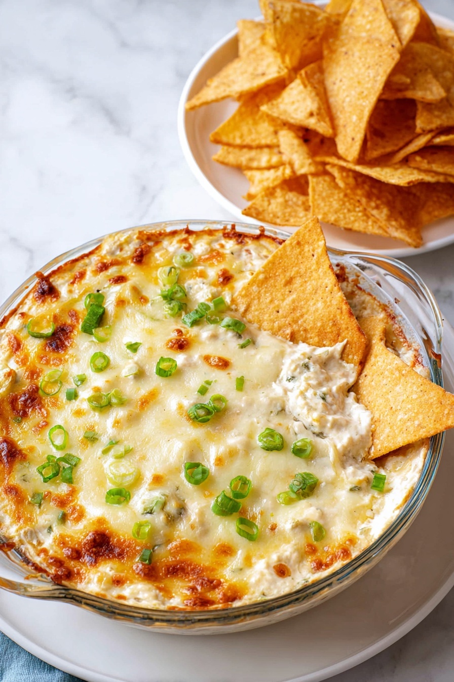 A clear round glass dish filled with a creamy baked dip that has a golden brown melted cheese top layer, slightly browned and bubbly around the edges, sprinkled with small bright green chopped scallions, with several triangular light brown crispy chips dipped into the creamy white layer under the cheese; next to the dish is a white plate stacked with more crispy triangular chips on a white marbled surface. photo taken with an iphone --ar 2:3 --v 7