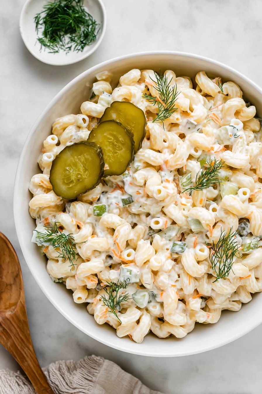 A white bowl holds a creamy pasta salad made of short, twisted macaroni coated in a white dressing with visible small herb bits. Mixed in are small pieces of green pickles and thin orange strands, likely carrots. On top of the salad are several green pickle slices and sprigs of fresh dill, adding a touch of color and texture. The bowl sits on a white marbled surface next to a small white dish with more dill and a wooden spoon nearby. photo taken with an iphone --ar 2:3 --v 7