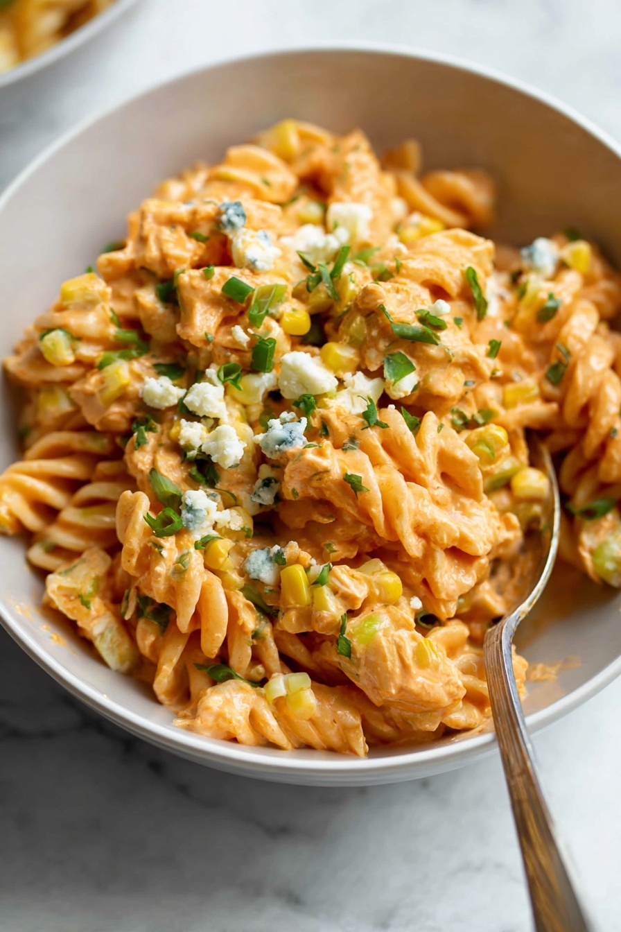 A close-up of a white bowl filled with orange-colored rotini pasta covered in a creamy sauce. The pasta is mixed with small yellow corn pieces and green chopped herbs scattered on top. There are also small white cheese crumbles spread over the dish, with some green herbs sprinkled on the cheese. A spoon rests inside the bowl on the right side, and the background shows a white marbled surface with a blurred hint of another dish. photo taken with an iphone --ar 2:3 --v 7