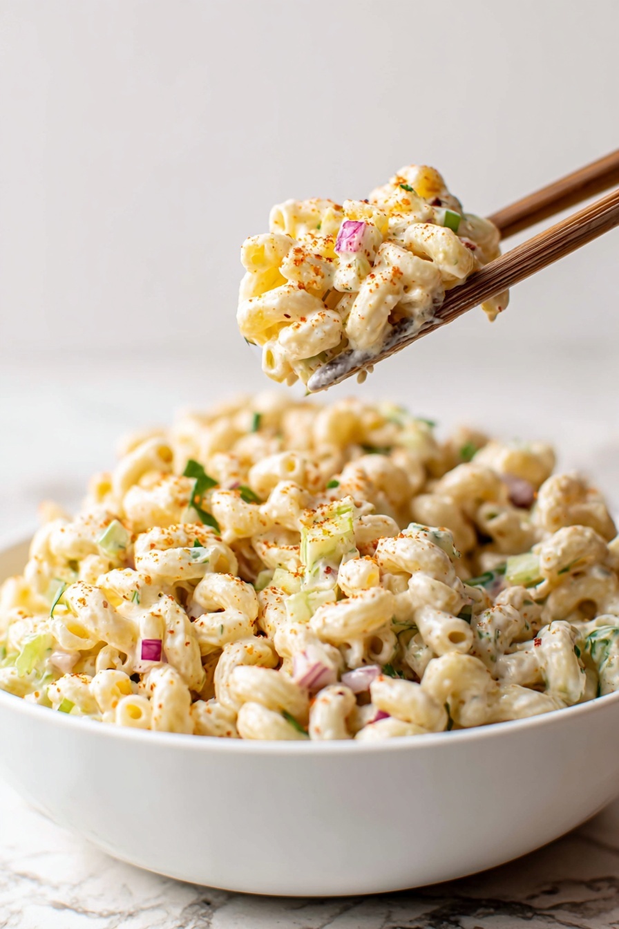 A white bowl filled with creamy macaroni salad that has small pieces of celery, finely chopped herbs, and bits of red onion mixed throughout, creating layers of pale cream, light green, and small red specks; a pair of wooden chopsticks holds a scoop of the salad above the bowl, showing the curly pasta coated in a smooth white dressing with some light orange seasoning dusted lightly on top; everything is set against a white marbled textured surface and background. photo taken with an iphone --ar 2:3 --v 7