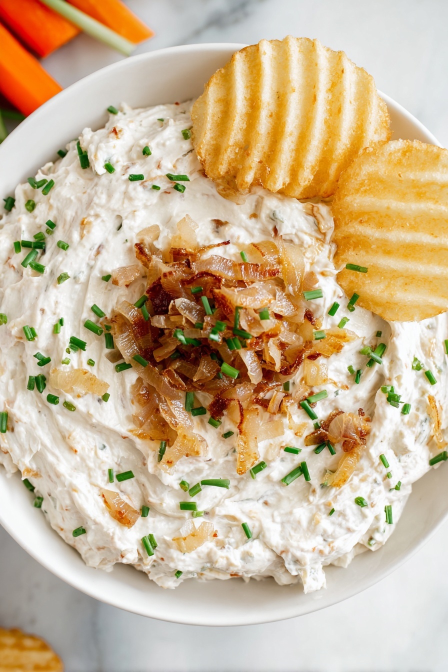A white bowl holds a creamy, thick dip spread evenly with a soft, slightly chunky texture. The base layer is white with small bits of onion mixed in. A ring of lightly browned cooked onions is placed on top, concentrated on one side of the bowl. Bright green chopped chives are sprinkled over the dip and onions, adding small pops of color. Two ridged potato chips rest on the dip near the cooked onions. The bowl sits on a white marbled surface with some carrot sticks partially visible at the top left corner. Photo taken with an iphone --ar 2:3 --v 7
