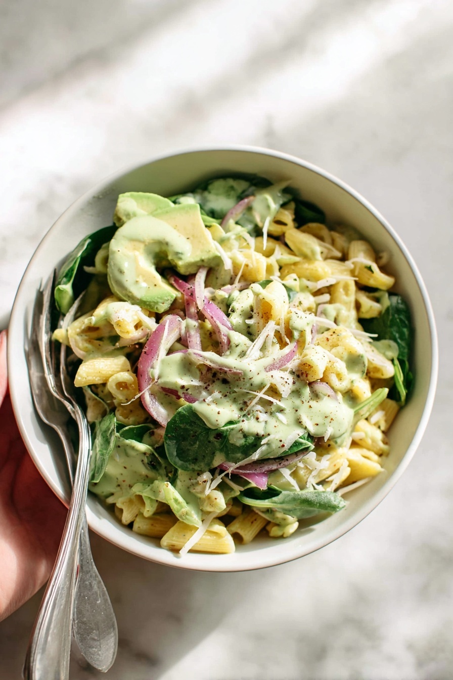 The image shows a white bowl filled with a pasta salad made of three main layers. The bottom layer is short pasta tubes in pale yellow color, mixed throughout with pieces of green leafy spinach. The middle layer has thin slices of purple-red onion and light green strips of avocado, with some pieces sprinkled with finely grated white cheese. The top layer is a creamy light green sauce drizzled over everything, adding a glossy texture. A woman's hand holds a silver spoon and fork resting on the side of the bowl. The bowl is placed on a white marbled surface with soft natural light creating gentle shadows. photo taken with an iphone --ar 2:3 --v 7
