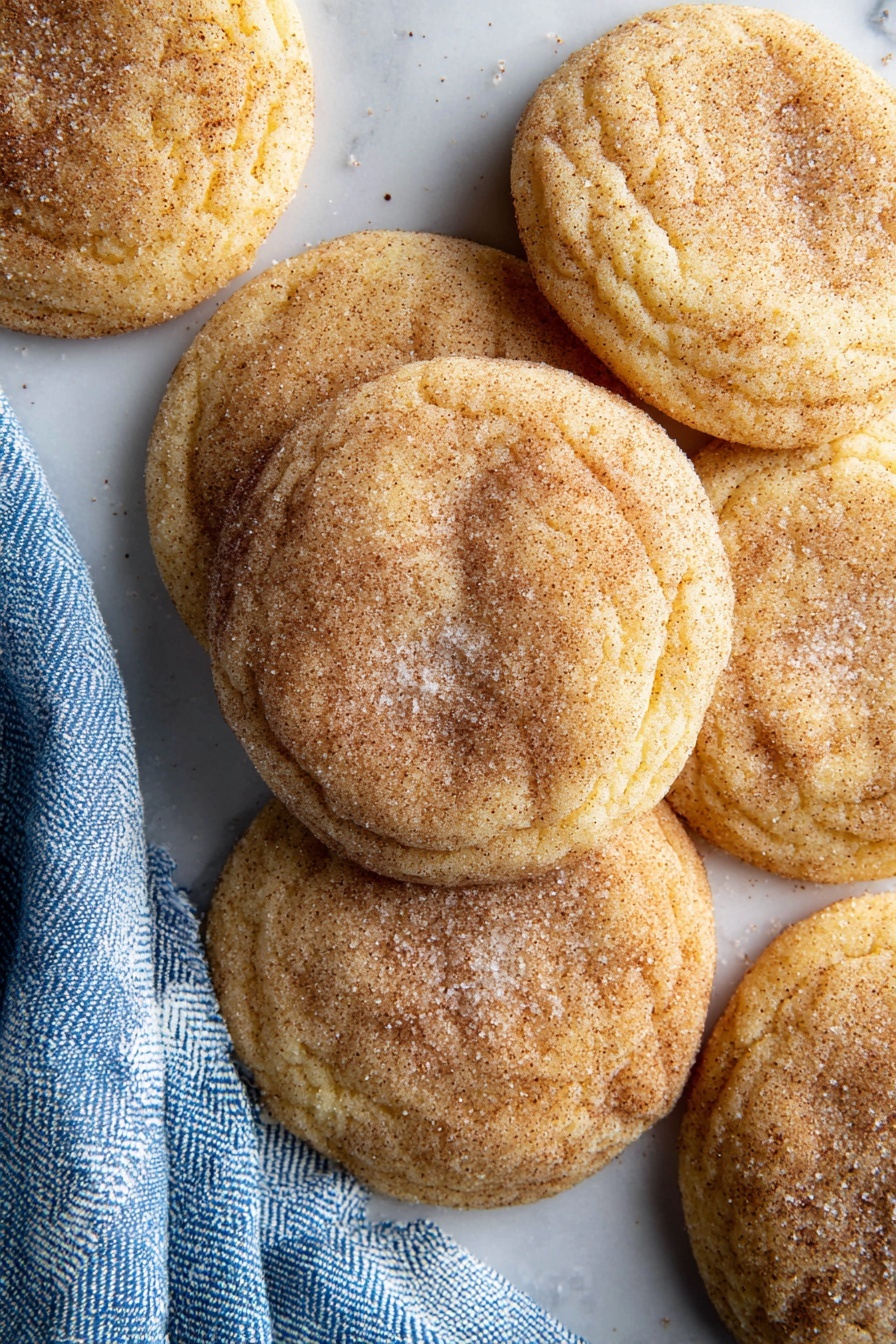 A close-up view of six round cookies arranged on a white marbled surface. The cookies have a golden-brown color with a soft texture, and they are sprinkled lightly with white sugar and cinnamon powder on top, giving a speckled effect. The edges of the cookies show slight wrinkles, indicating a soft and chewy center. A blue and white checkered cloth is placed near the lower left corner, adding a soft fabric texture to the scene. The lighting highlights the baked texture and sugar dusting, making the cookies look warm and inviting. Photo taken with an iphone --ar 2:3 --v 7
