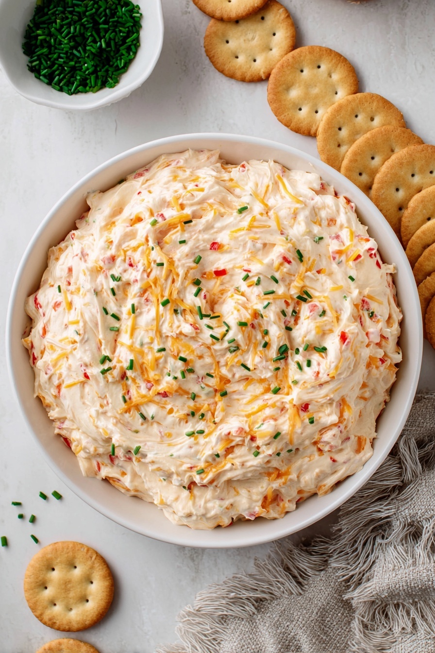 A close-up view of a white bowl filled with a creamy, thick cheese spread mixed with visible small bits of orange shredded cheese and tiny red pieces, likely peppers, all swirled together. The spread is topped with scattered small green chives on its surface, adding a pop of color. Around the bowl are round light brown crackers arranged in a slight fan shape on a white marbled surface. In the top left corner, a small white bowl holds more finely chopped green chives, with some chives scattered nearby. A textured light gray fringed cloth is partially visible on the right side. The photo taken with an iphone --ar 2:3 --v 7