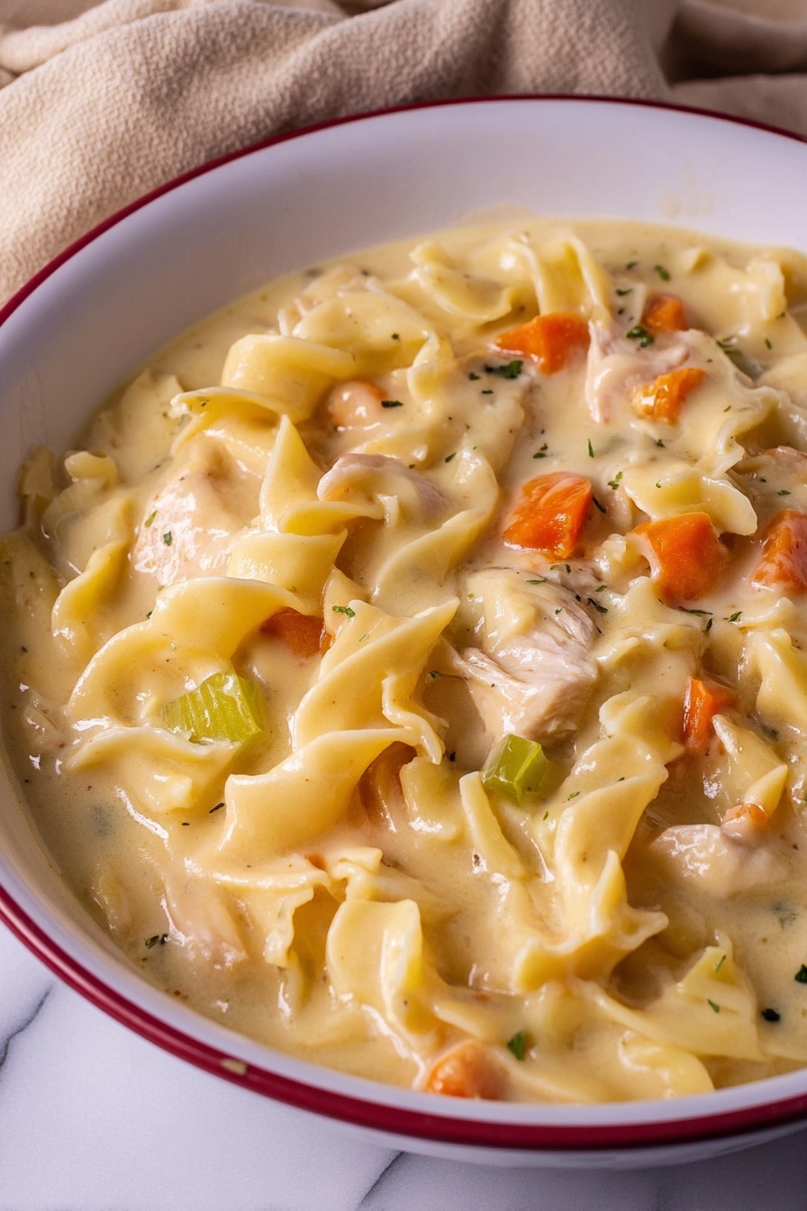 A close-up view of a white bowl with a red ring around the edge filled with creamy chicken and vegetable noodle soup. The dish has a thick, pale yellow creamy broth covering soft wide egg noodles that have wavy edges. In the soup, there are small pieces of cooked chicken and diced vegetables including bright orange carrots and light green celery. The texture looks smooth and hearty, with the noodles and vegetables mixed evenly through the creamy sauce. The bowl sits on a white marbled surface, with a beige cloth partially visible underneath. photo taken with an iphone --ar 2:3 --v 7