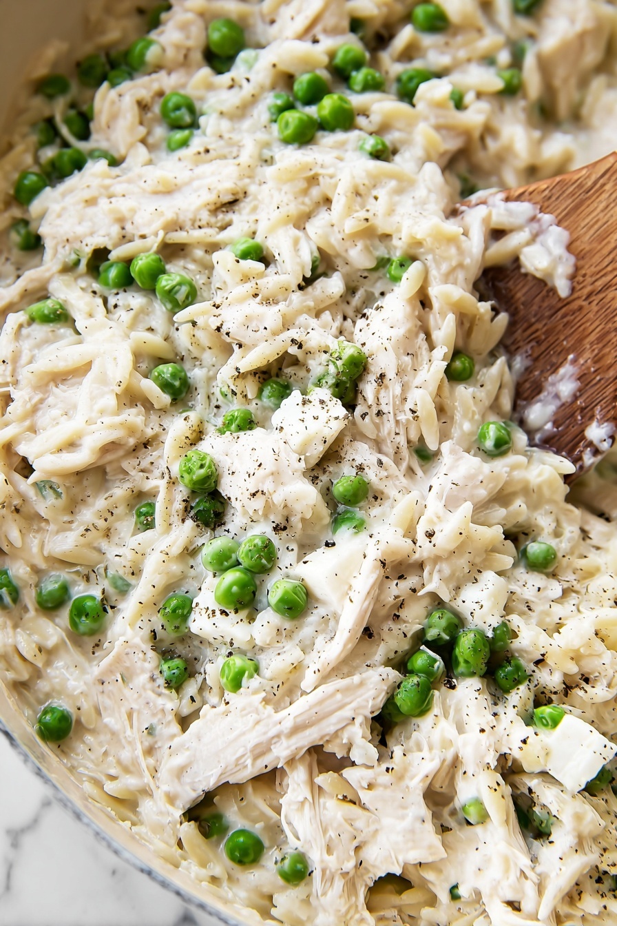 The image shows a creamy dish with three main layers. The base layer is made of soft white orzo pasta mixed with shredded white chicken, both covered in a thick white sauce. Scattered evenly throughout the dish are bright green peas, adding a fresh pop of color. On top, there are chunks of white cheese sprinkled with black pepper for texture. A wooden spoon is partly visible on the right side, stirring the dish. The background has a white marbled texture. photo taken with an iphone --ar 2:3 --v 7