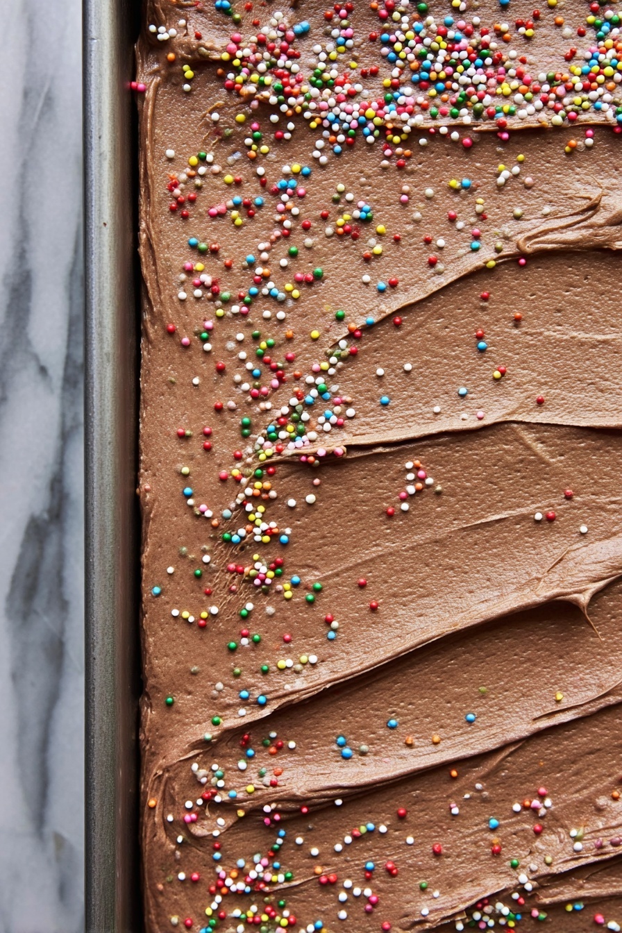 A close-up view of a rectangular cake with one thick layer covered in smooth, creamy chocolate frosting spread unevenly with visible swirls and texture. The top is decorated with small, round, colorful sprinkles scattered unevenly across the surface. The cake sits in a metal baking tray, and the background shows a white marbled texture. Photo taken with an iphone --ar 2:3 --v 7