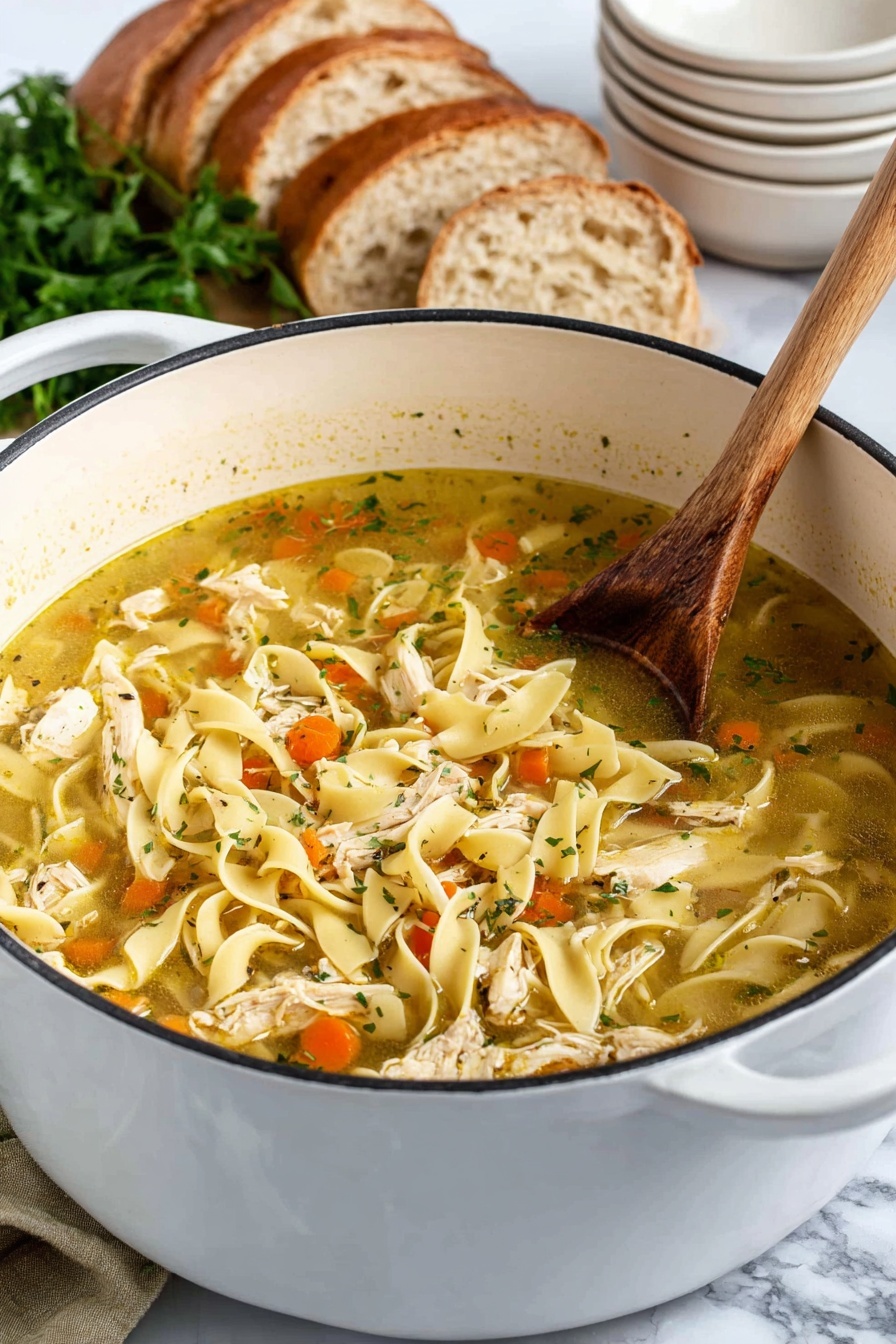 The image shows a white bowl filled with chicken noodle soup on a white marbled surface. The soup has clear broth with visible layers including wide, flat pale yellow noodles, thin slices of cooked chicken, bright orange carrot rounds, and light green celery pieces mixed with small bits of herbs. A spoon lifts a portion of the soup from the bowl, showing a detailed mix of these ingredients. In the background is a large pot filled with more of the same soup and two pieces of bread on the surface nearby. Photo taken with an iphone --ar 2:3 --v 7
